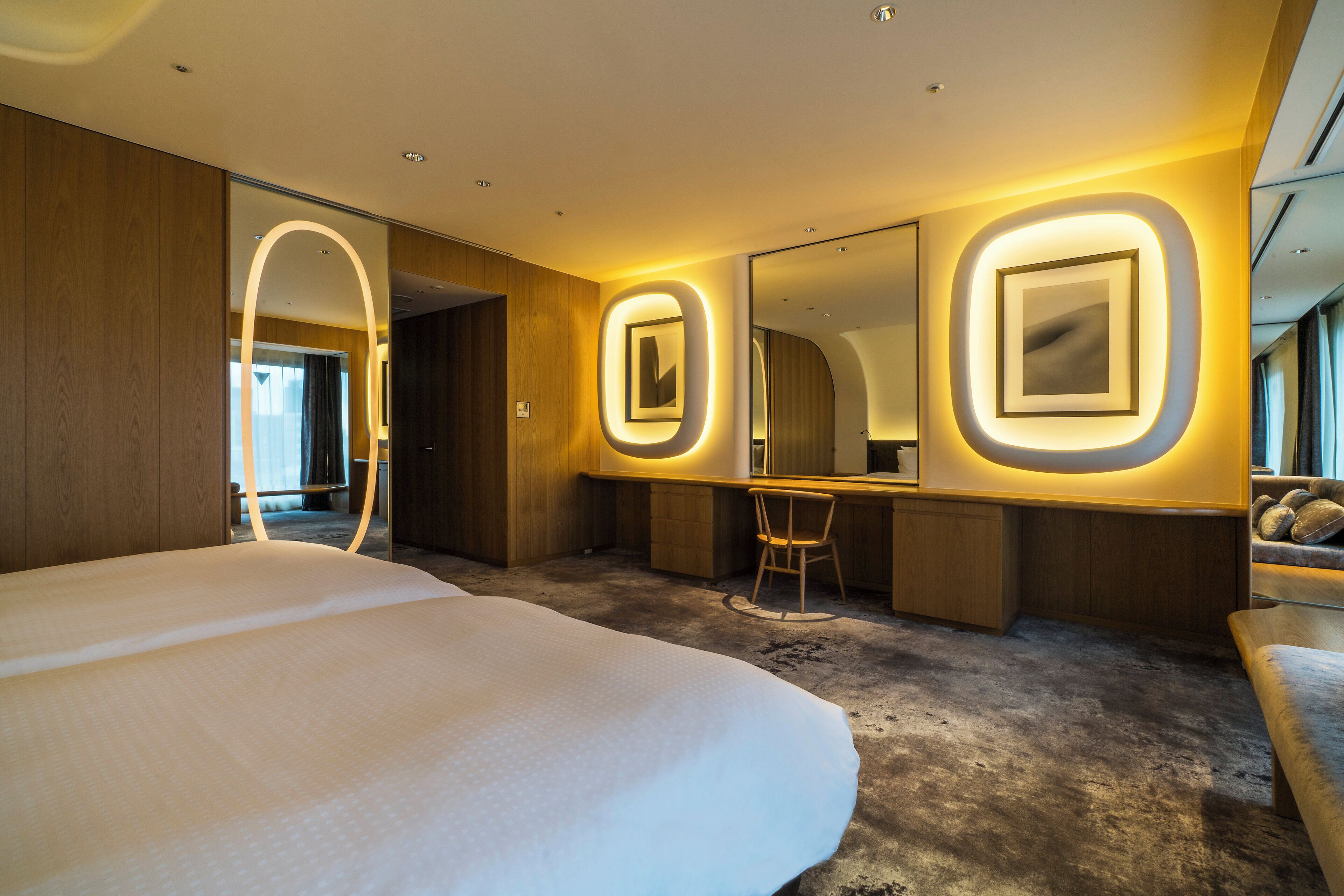 Una habitación de hotel moderna con una cama grande, un escritorio y una silla. Hay dos espejos montados en la pared con marcos iluminados.