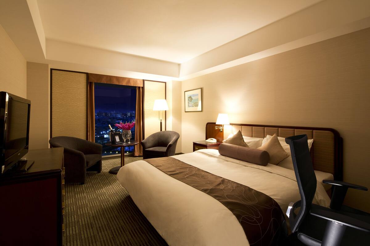 A hotel room with a large bed, a desk with a TV, a chair, a nightstand with a lamp, and a window with a city view.