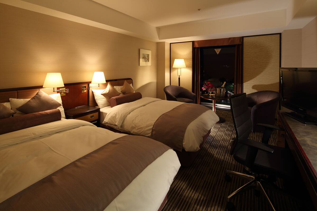 A hotel room with two beds, a desk, a chair, and a TV.
