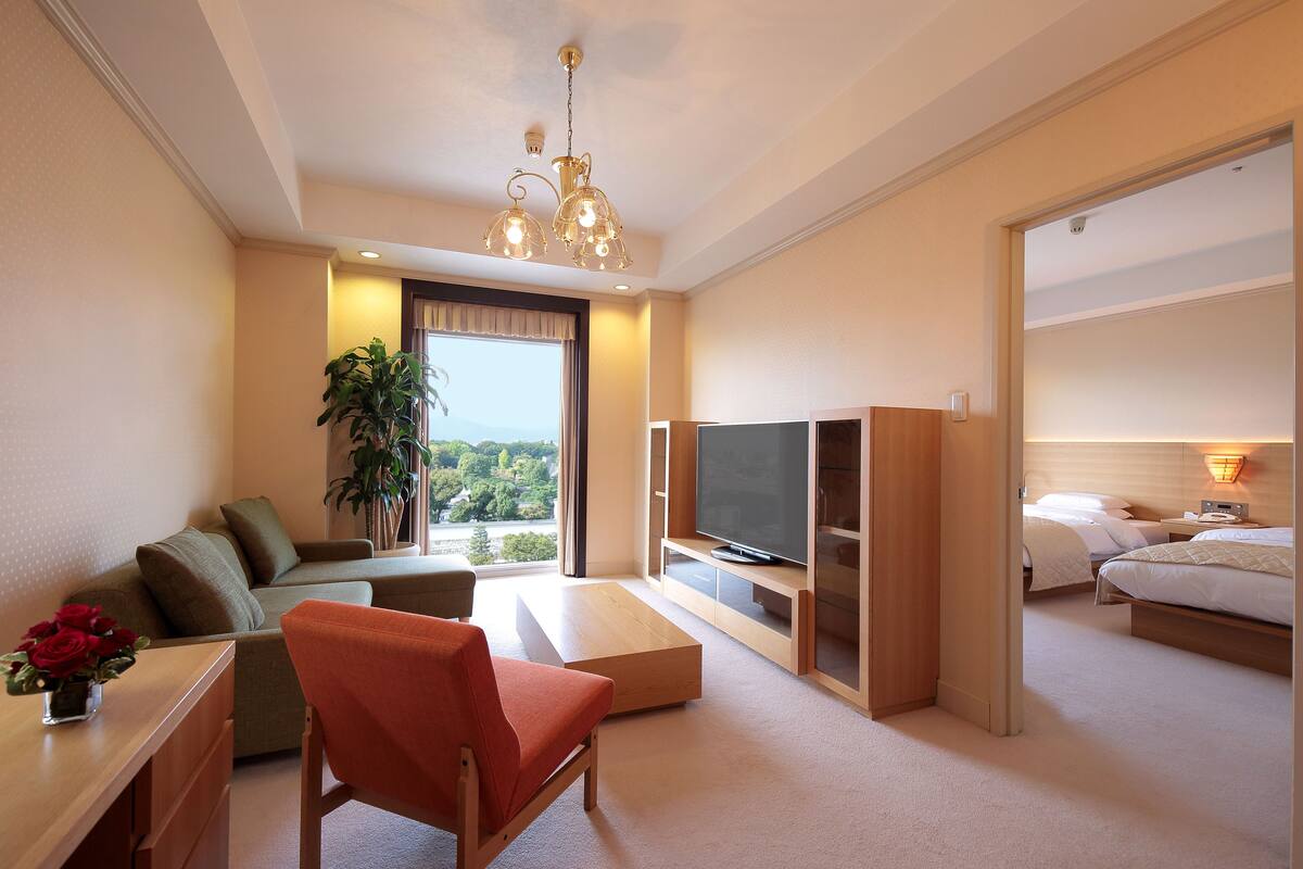 A hotel room with a sofa, a red chair, a TV, a bed, and a large window with a view.