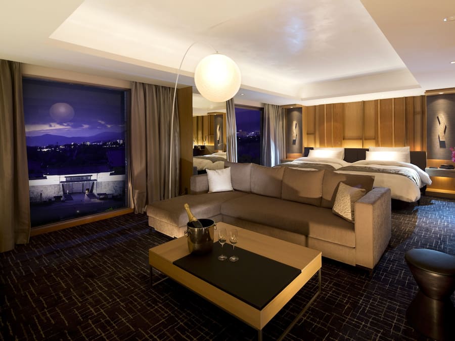 Quarto de hotel moderno com uma cama grande, um sofá, uma mesa de centro e vista para a cidade à noite.