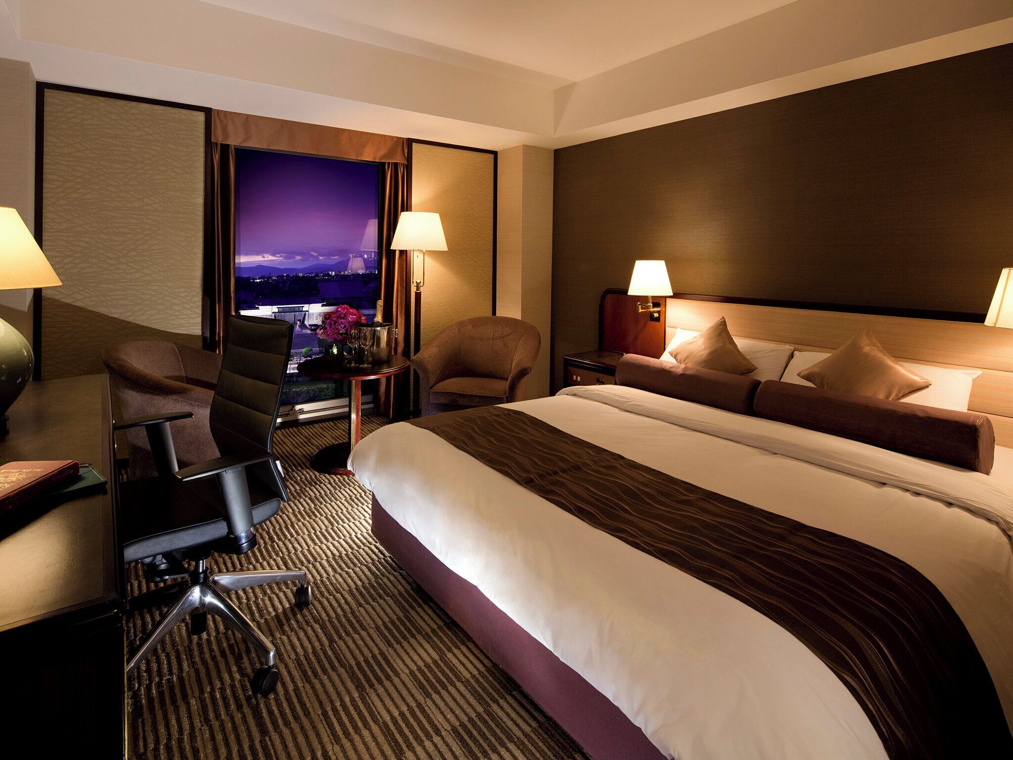 Ein Hotelzimmer mit einem großen Bett, zwei Sesseln, einem Schreibtisch mit Stuhl und Blick auf die Stadt.