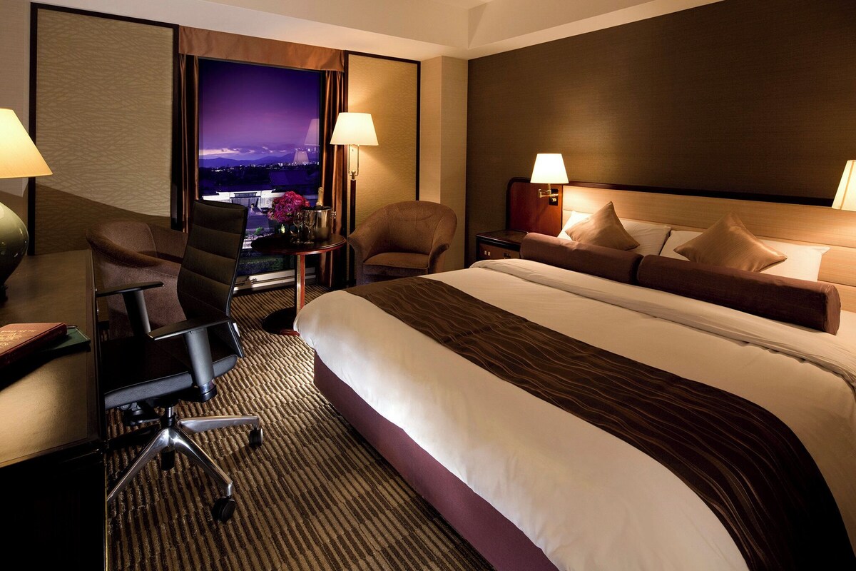Une chambre d’hôtel avec un grand lit, deux fauteuils, un bureau avec une chaise et une vue sur le paysage urbain.