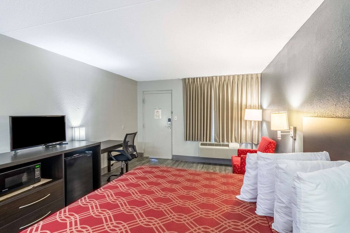 Habitación de hotel con cama, escritorio con televisor, refrigerador pequeño, silla y ventana con cortinas.
