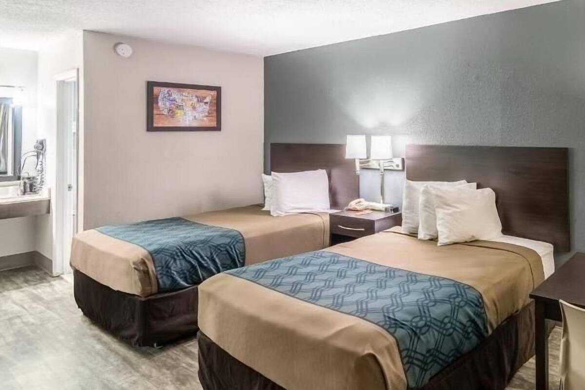 Habitación de hotel con dos camas, un escritorio, una silla, una mesita de noche y una lámpara fijada a la pared.