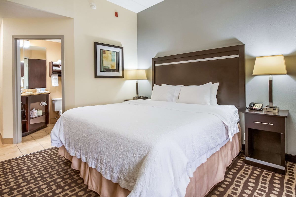 Quarto de hotel com uma cama grande, duas luminárias de cabeceira, mesa de cabeceira com telefone, quadro na parede e banheiro visível por uma porta aberta.