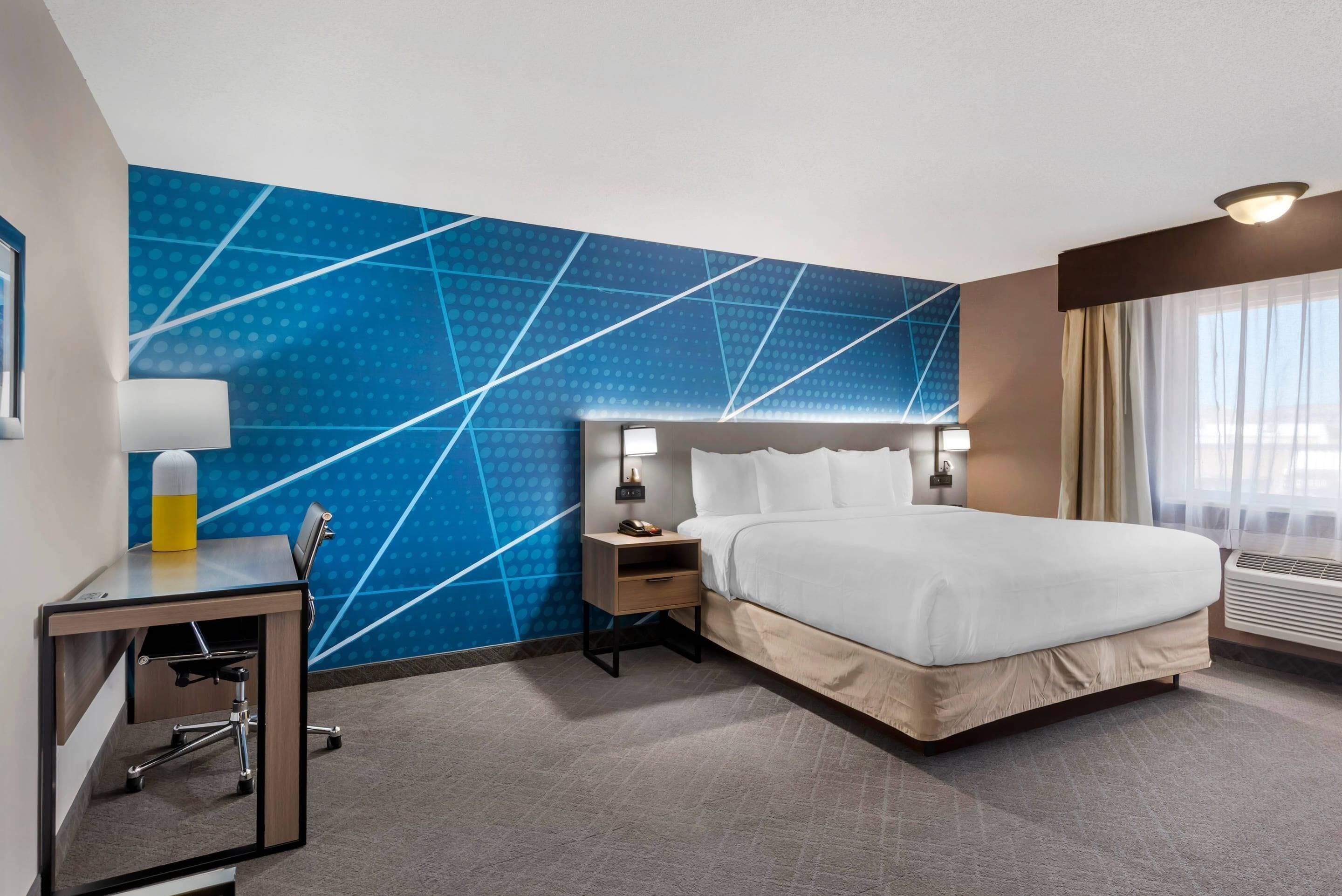 A hotel room with a bed, desk, chair, nightstand, lamp, and a blue patterned wall.