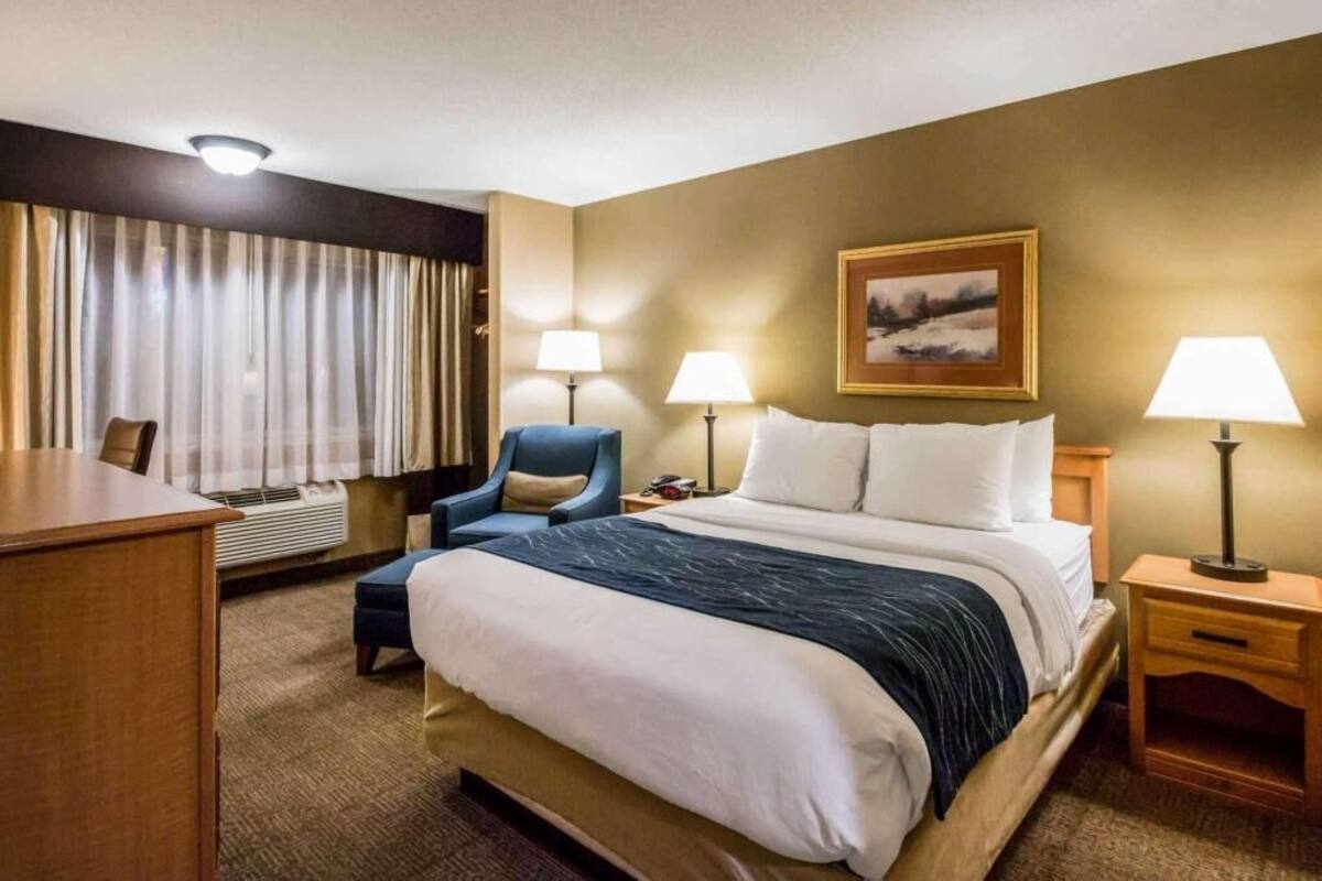 A hotel room with a bed, a desk, a chair, two bedside lamps, a framed picture on the wall, and a window with curtains.