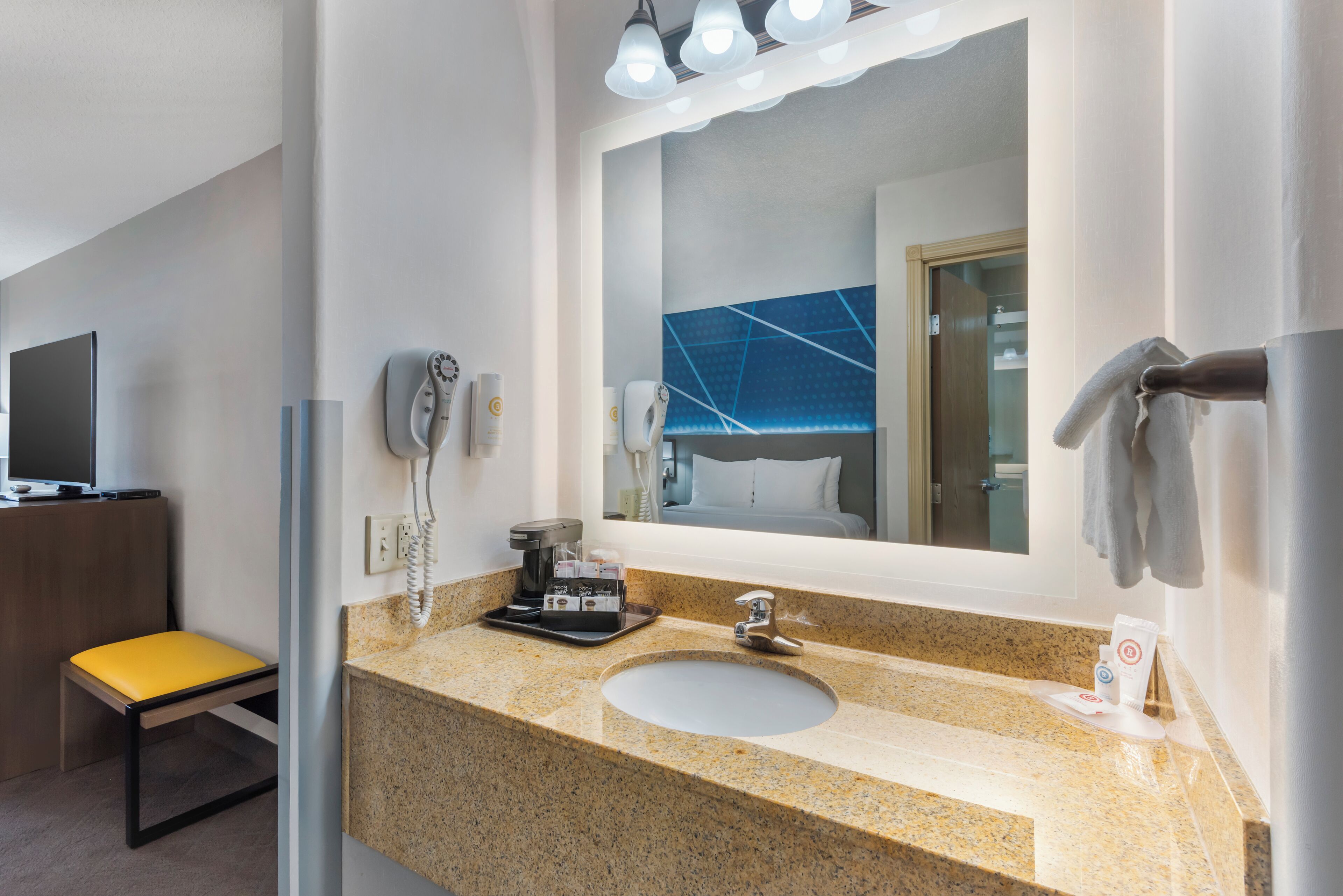 Un bagno d'hotel con un piano di lavoro in granito, un lavandino, uno specchio, un portasciugamani, un telefono e una piccola area salotto.
