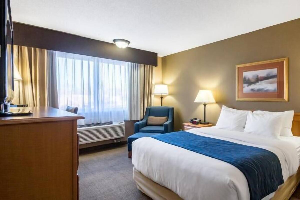 A hotel room with a bed, a desk, a chair, a nightstand, a lamp, and a painting on the wall.