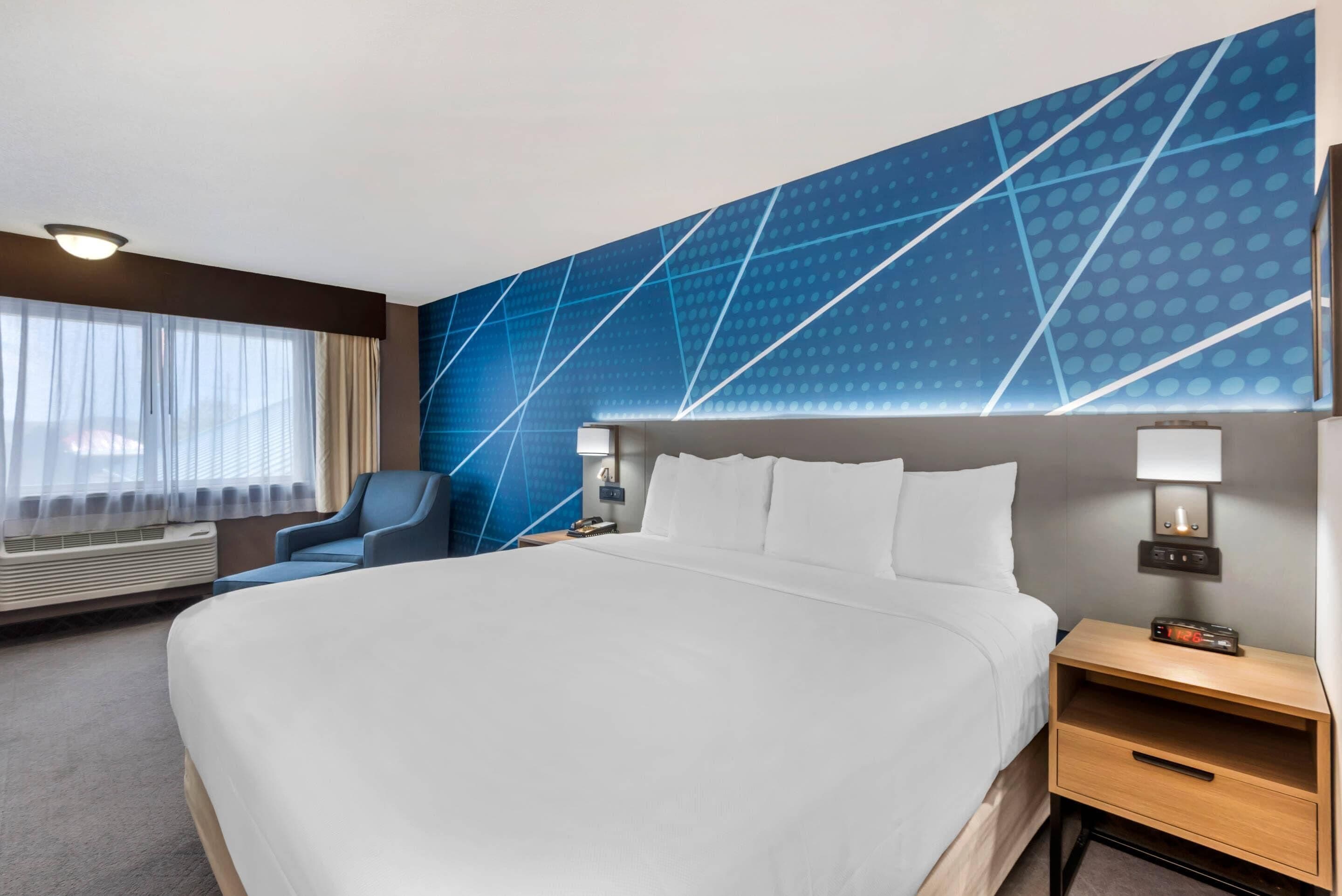 A hotel room with a large bed, a blue accent wall, a window with curtains, a chair, and a bedside table with a lamp.