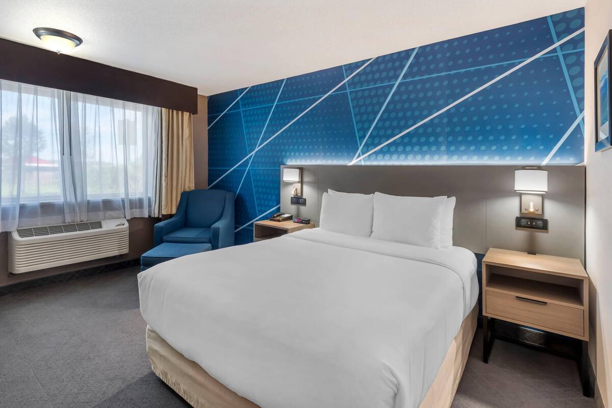 A hotel room with a large bed, a chair, two bedside tables with lamps, a window with sheer curtains, and a wall with a geometric pattern.