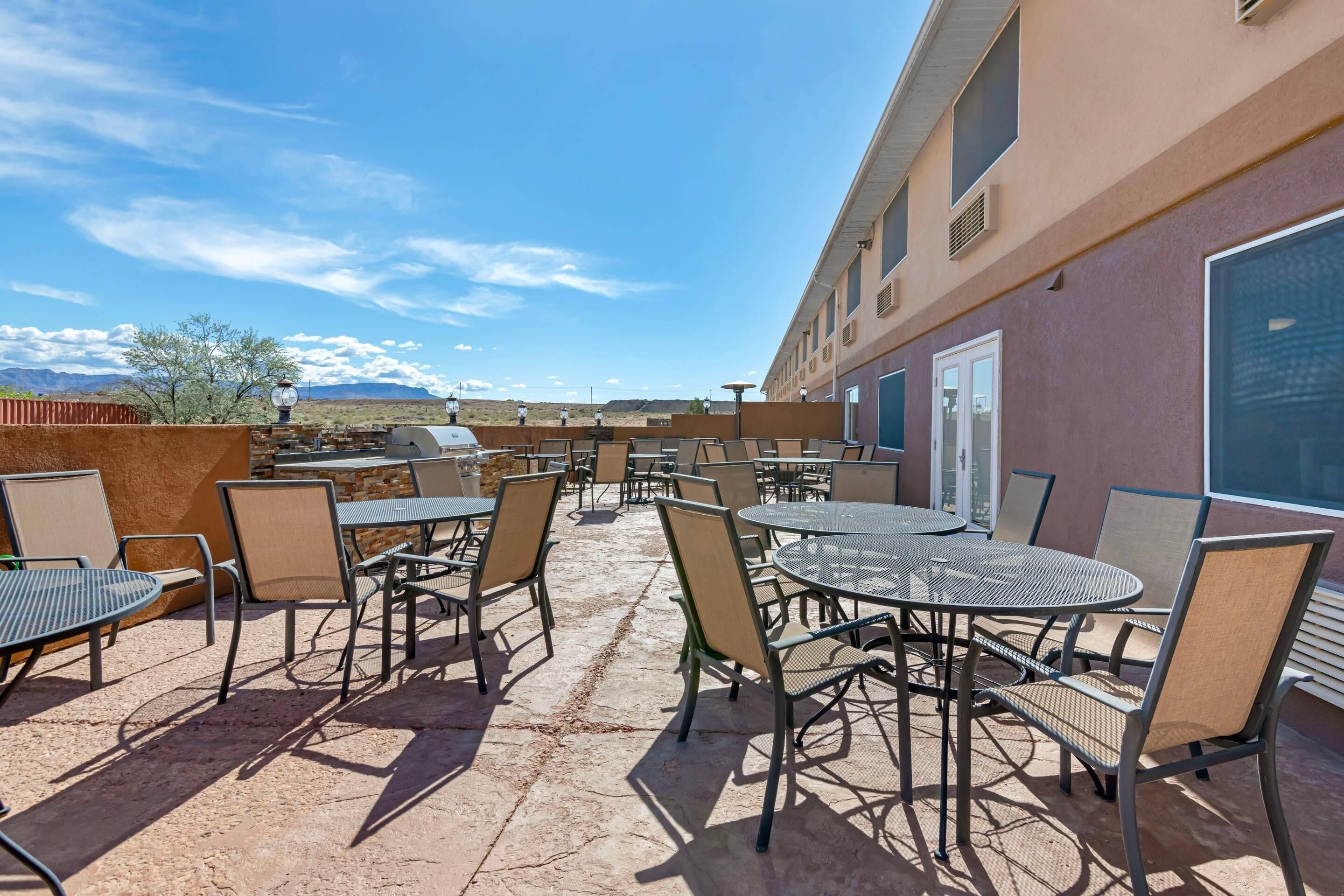 An outdoor patio with tables and chairs, a building with large windows, and a desert landscape in the background.