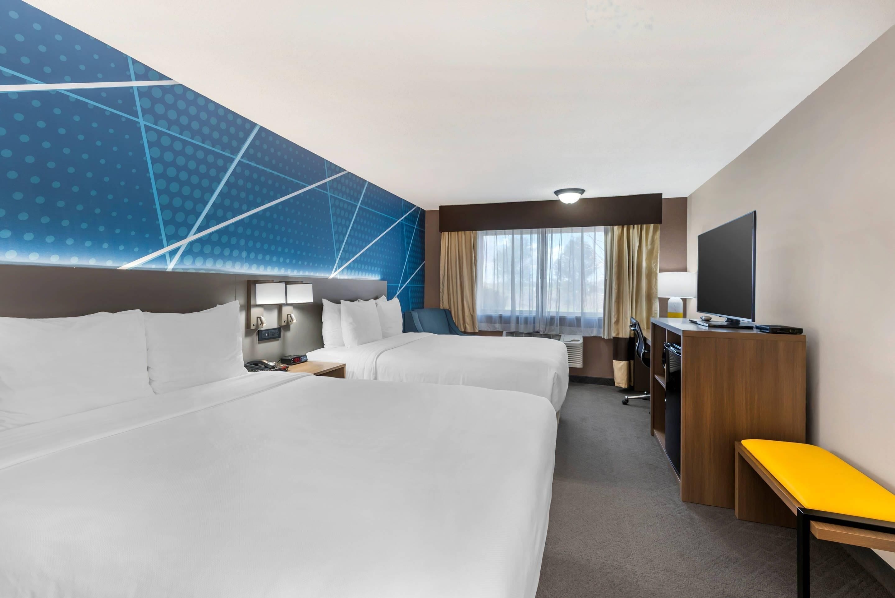 A hotel room with two beds, a desk, a TV, and a large window with curtains.