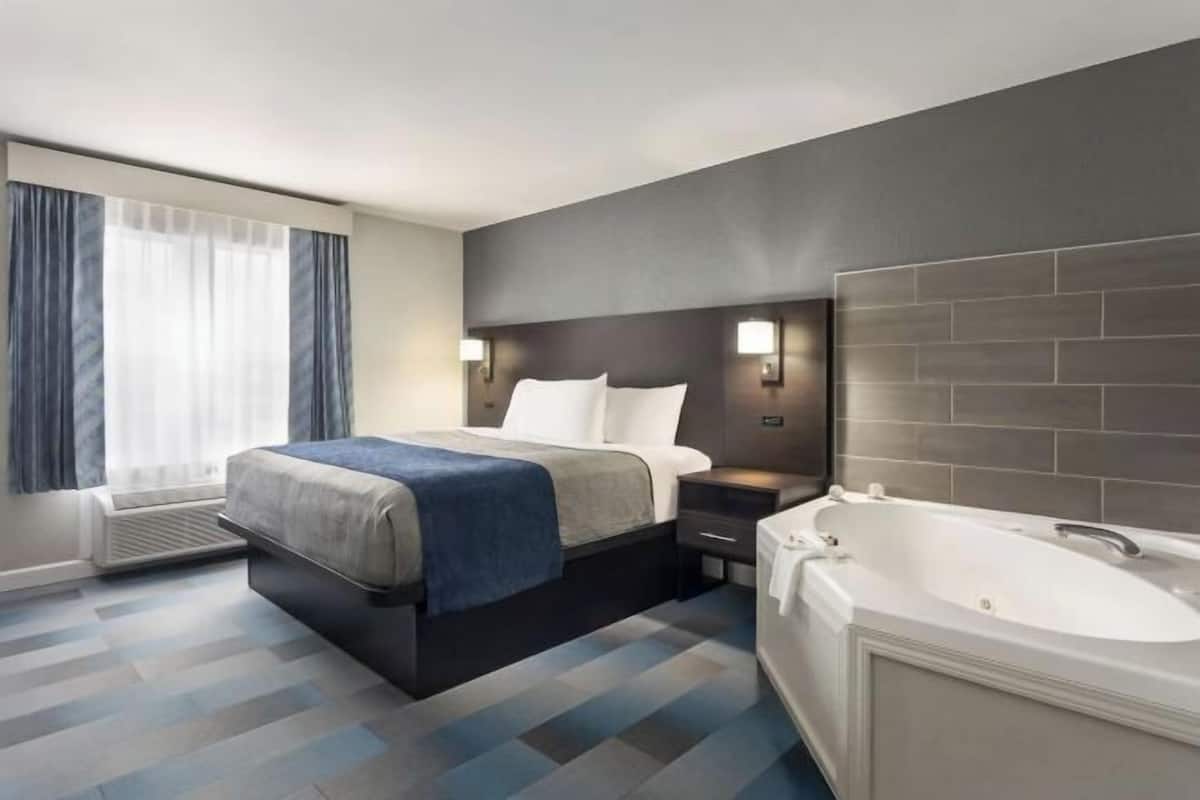 A modern hotel room with a bed, bedside table, lamp, and a large bathtub.