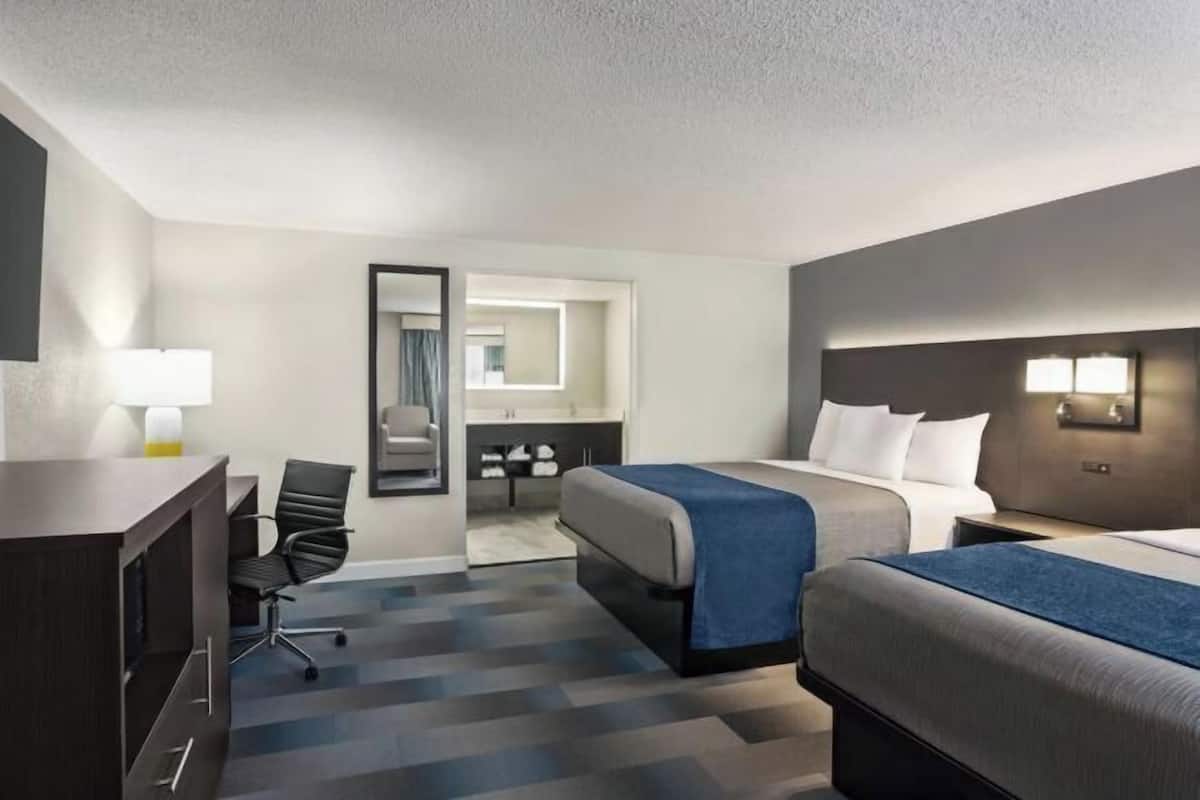 A hotel room with a desk, two beds, a chair, a mirror, and a bathroom visible through an open door.