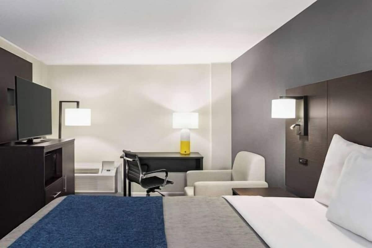 A hotel room with a bed, a desk, a chair, a TV, and two wall-mounted lamps.