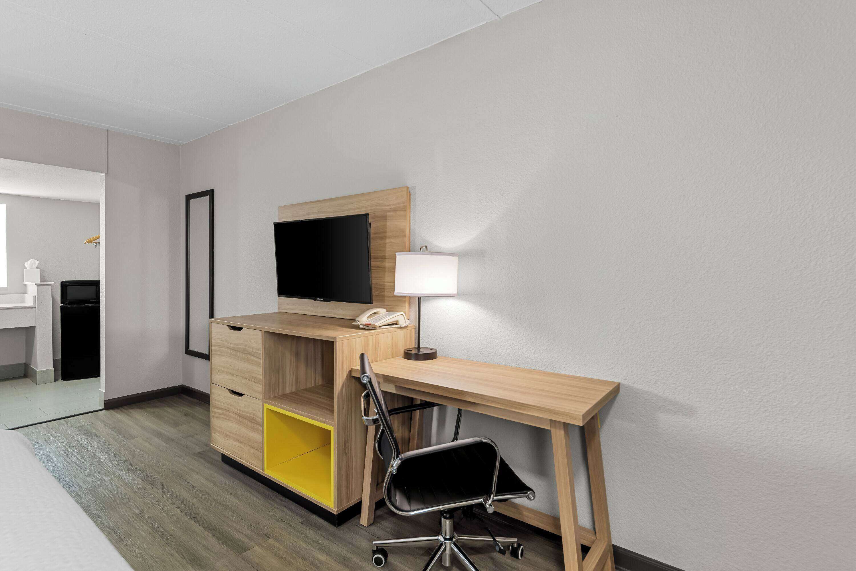 Una habitación de hotel con un escritorio de madera, una silla, un televisor montado en la pared, una mesita de noche y una cama.