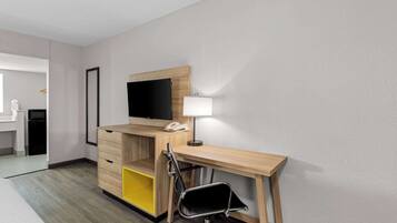 Una habitación de hotel con un escritorio de madera, una silla, un televisor montado en la pared, una mesita de noche y una cama.