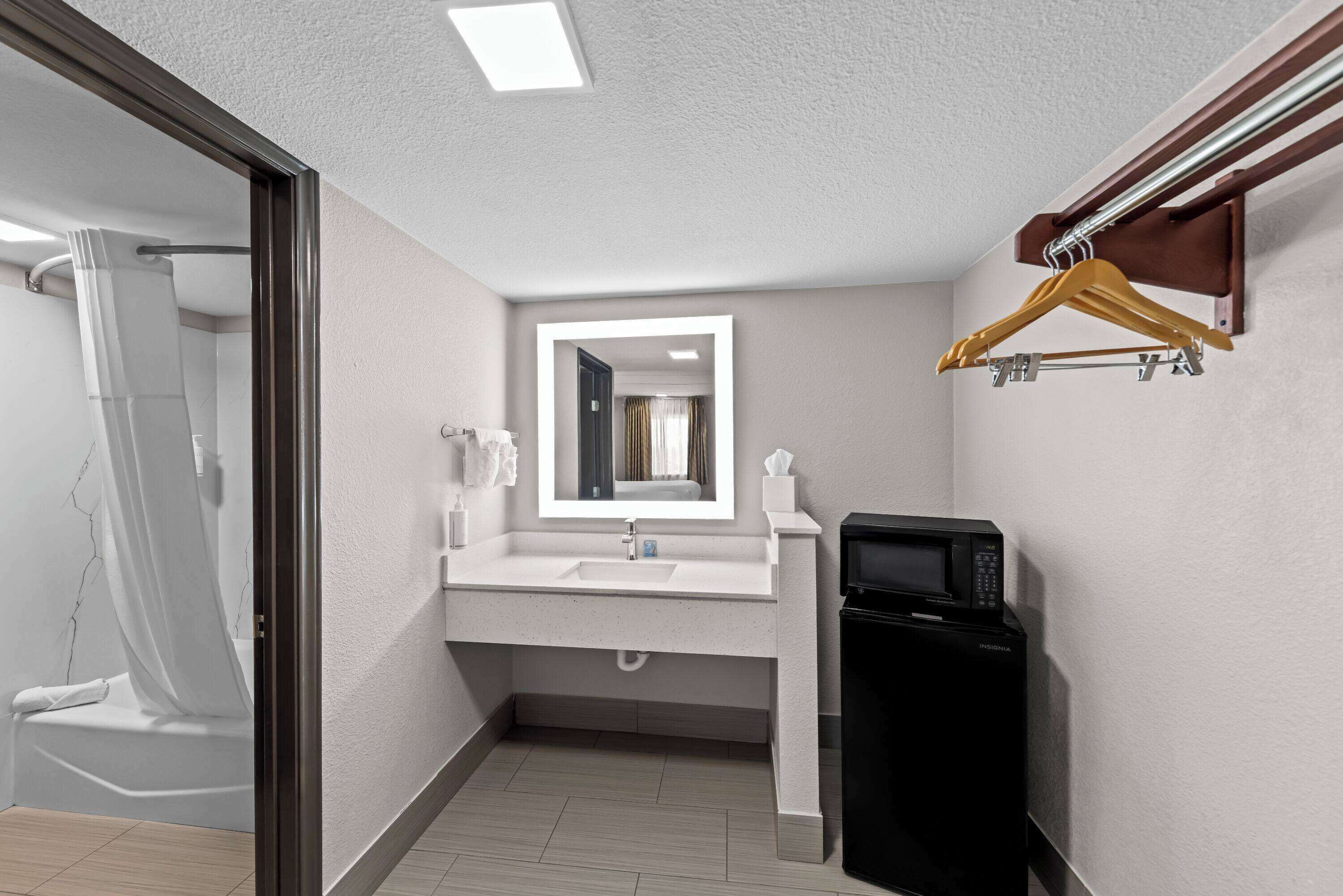 A modern bathroom with a white sink, a mirror, a black microwave, and a wooden hanger rack.