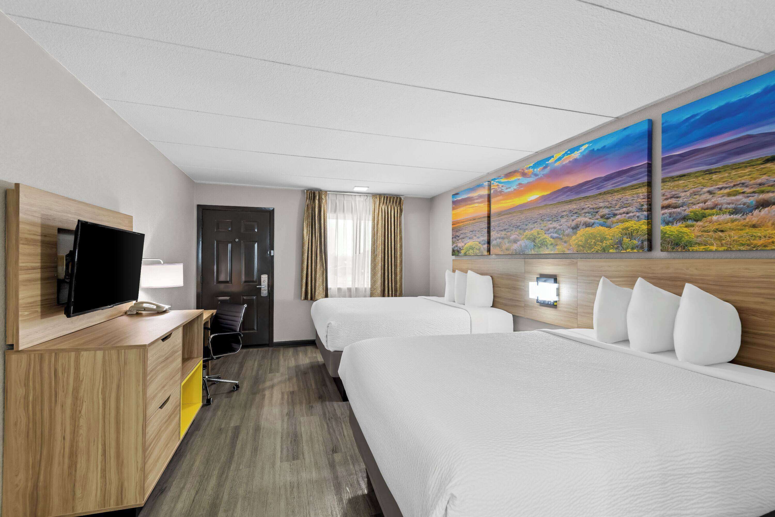 Quarto de hotel com duas camas, uma mesa de escritório com uma TV, uma luminária e um quadro de um pôr do sol.