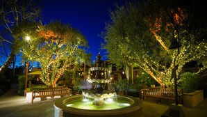 A nighttime outdoor area with a multi-tiered fountain, benches, and trees adorned with lights. - The Garland (North Hollywood)