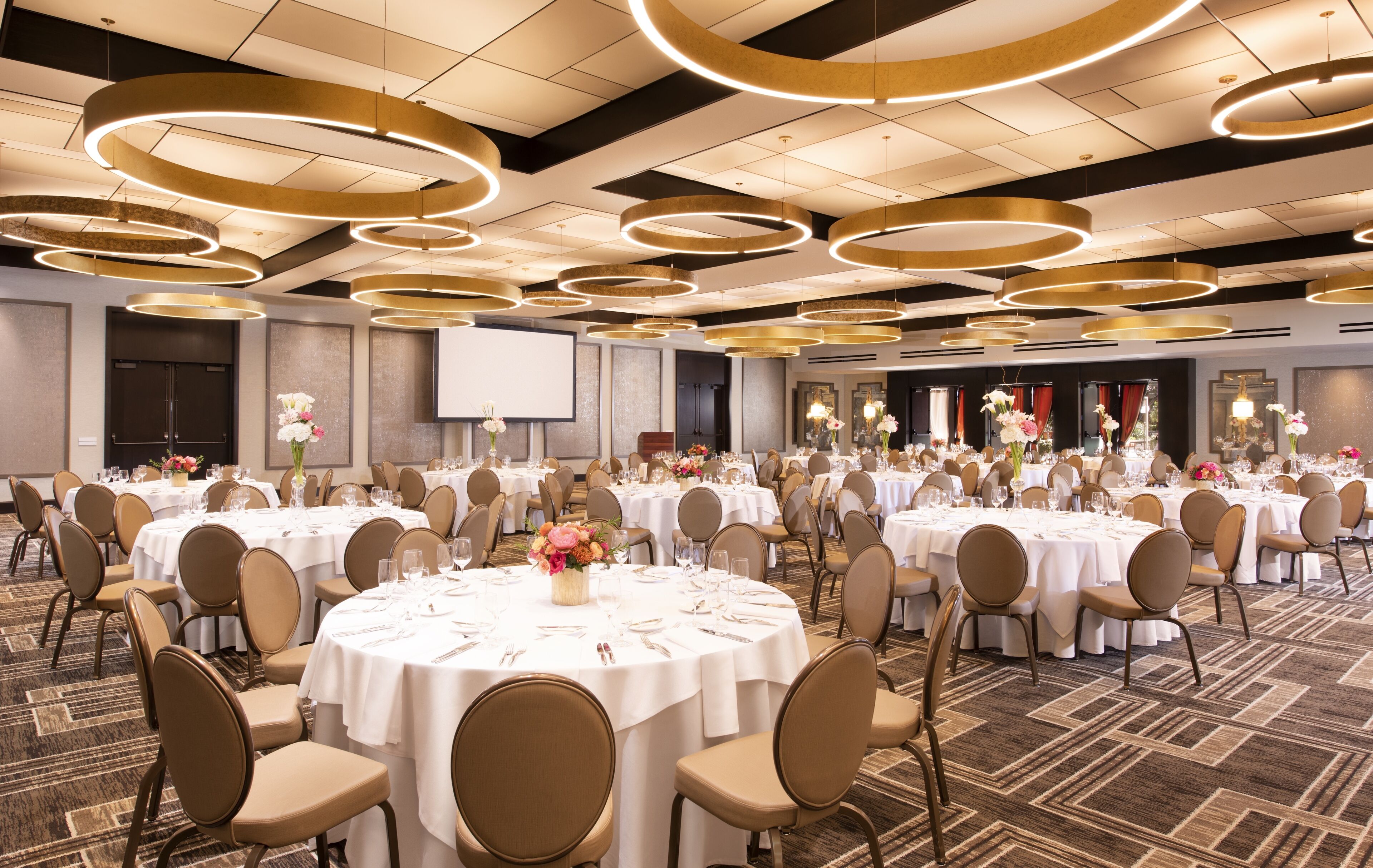 A large banquet hall with round tables, chairs, and a stage area.