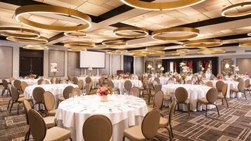 A large banquet hall with round tables, chairs, and a stage area.