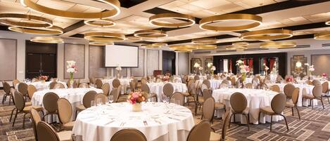 A large banquet hall with round tables, chairs, and a stage area.