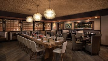 Un restaurant avec une longue table en bois, des chaises en osier et des lampes suspendues.