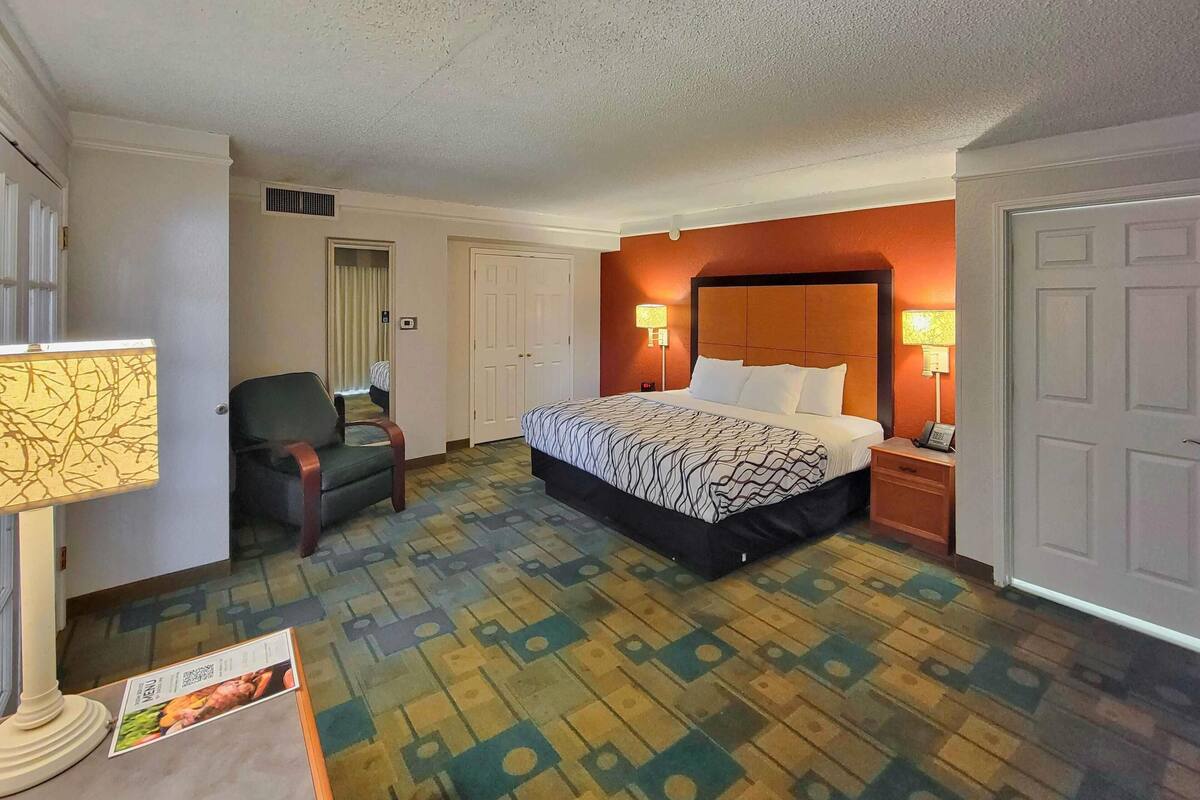 A hotel room with a bed, two bedside lamps, a chair, a desk with a lamp, and a wall-mounted air vent.