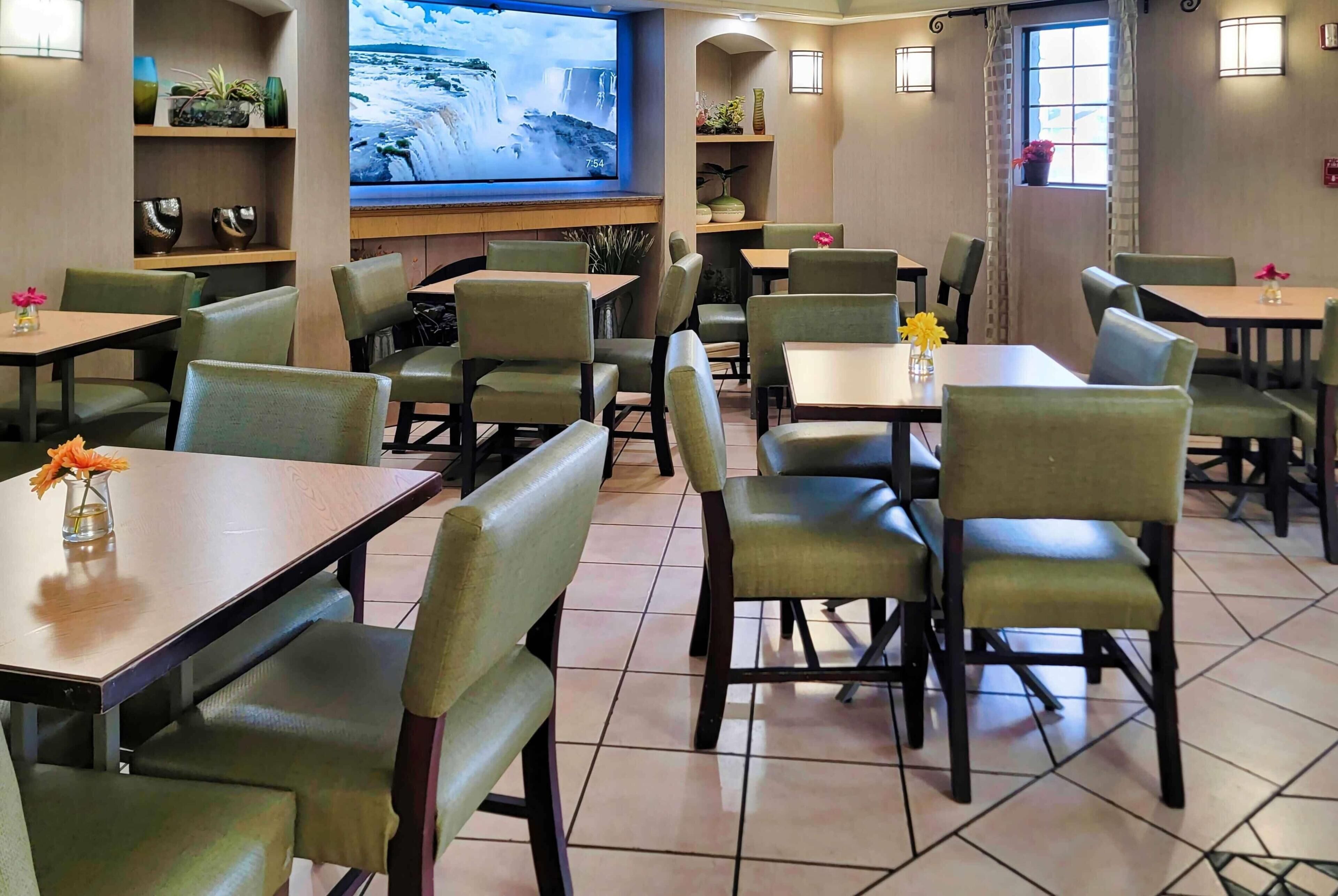 En restaurant med flere borde og stole, et stort skærmbillede af et vandfald, og reoler med dekorative genstande.