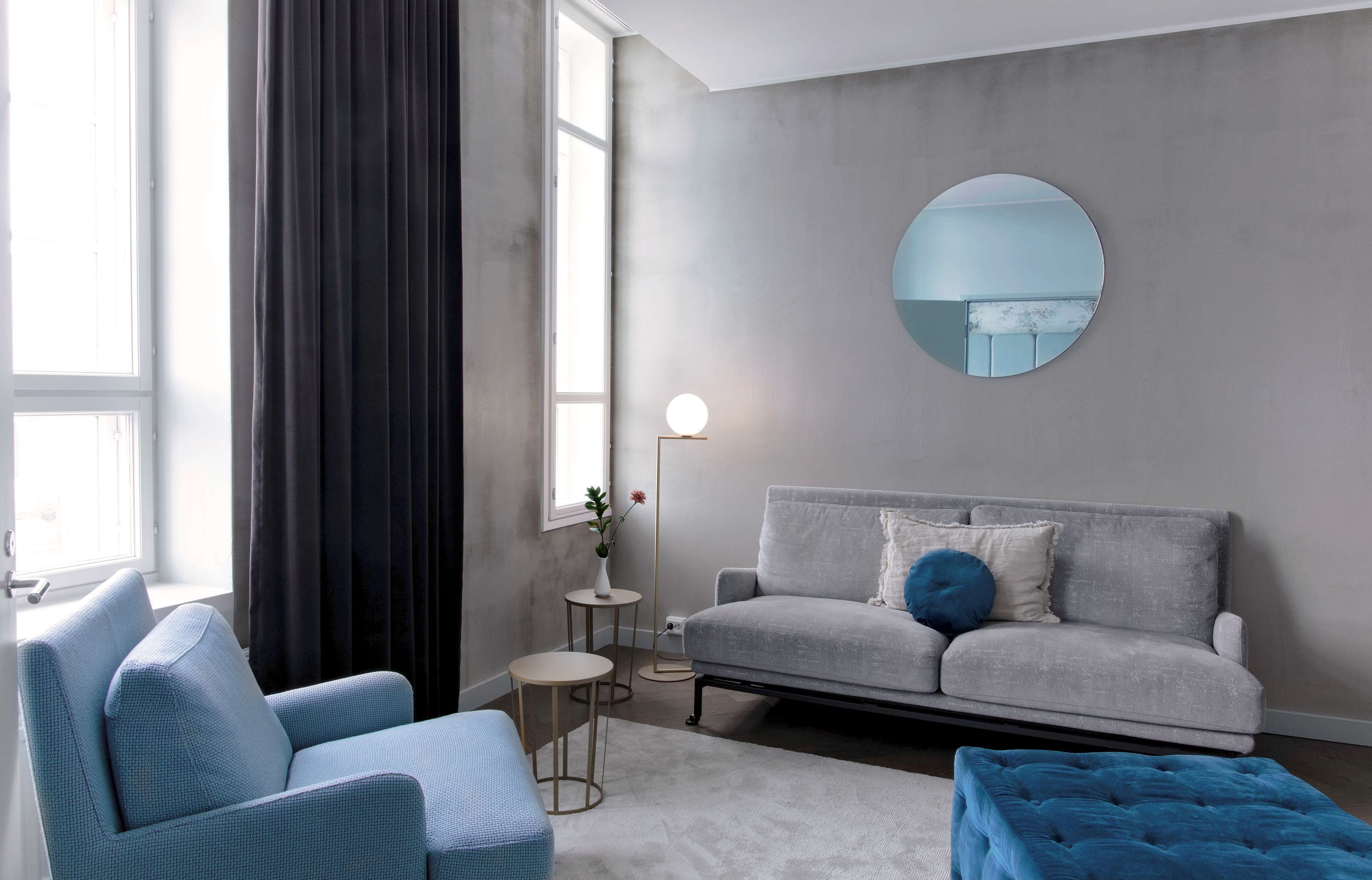 Una sala de estar moderna con un sofá gris, dos sillones, un espejo redondo y una mesita auxiliar con una lámpara.