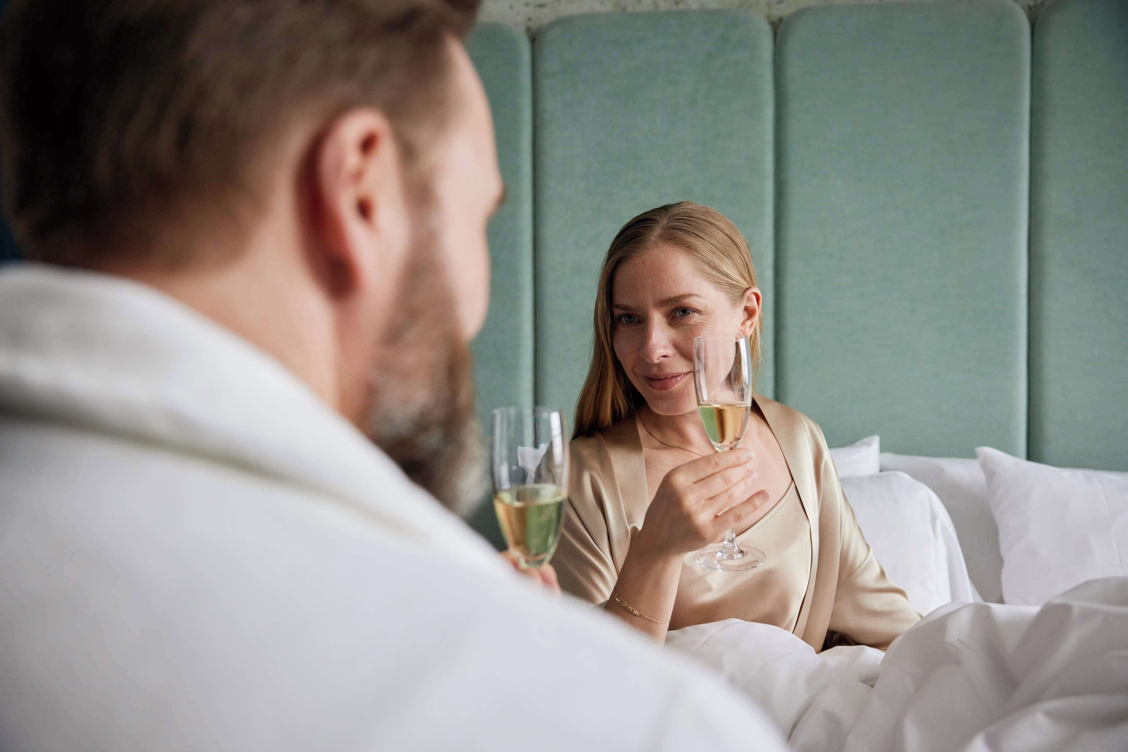 Eine Person in einem weißen Bademantel sitzt auf einem Bett mit einem grünen Kopfteil und hält ein Glas Weißwein.
