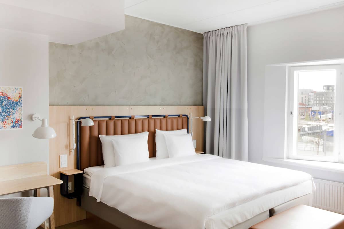 Moderni hotellihuone, jossa on suuri sänky, työpöytä ja ikkunasta avautuva näkymä kaupunkiin.