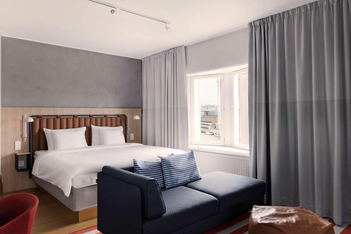 Quarto de hotel moderno com cama grande, sofá e janela com cortinas.
