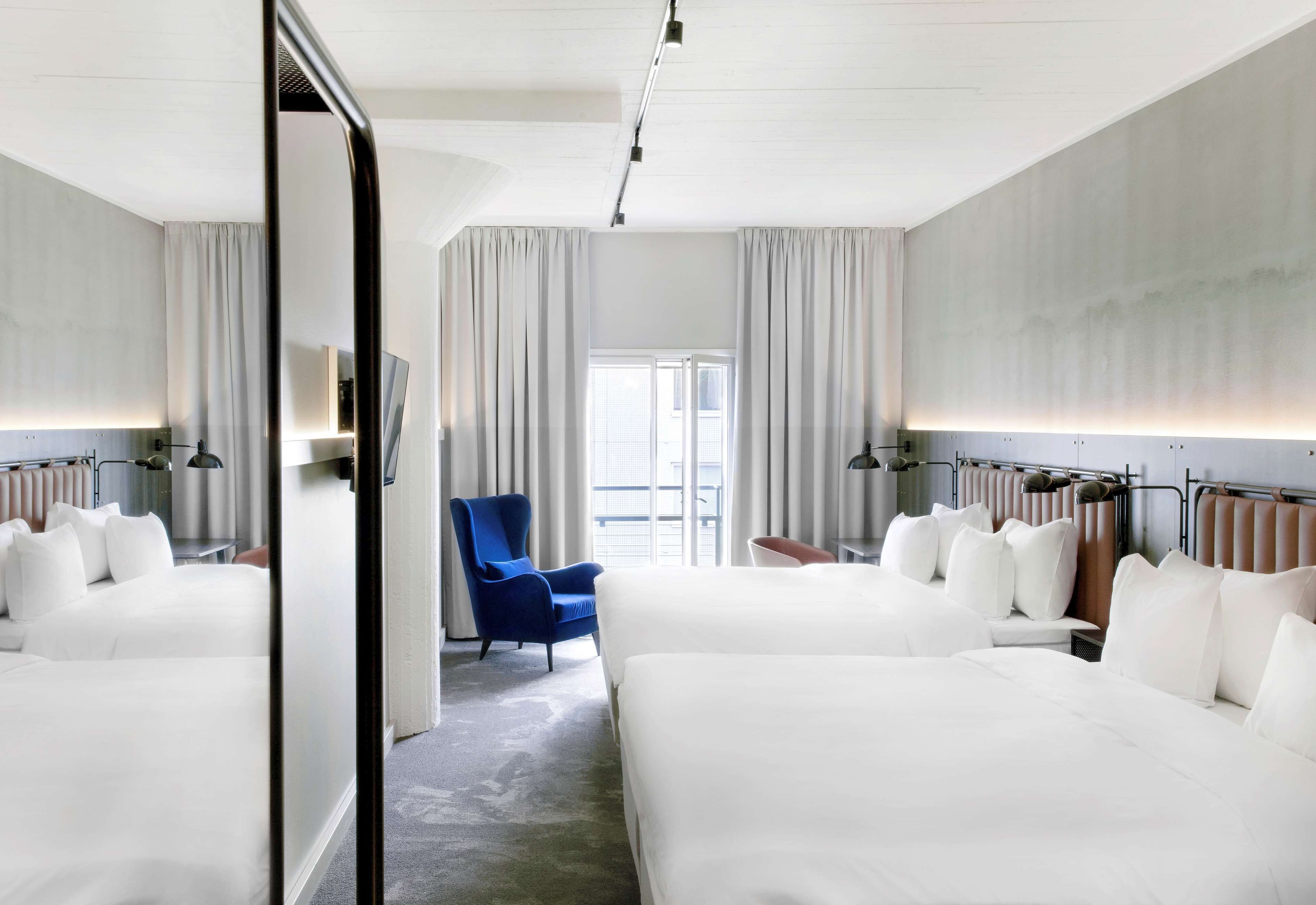 A hotel room with two beds, a blue armchair, and a view of the outdoors.