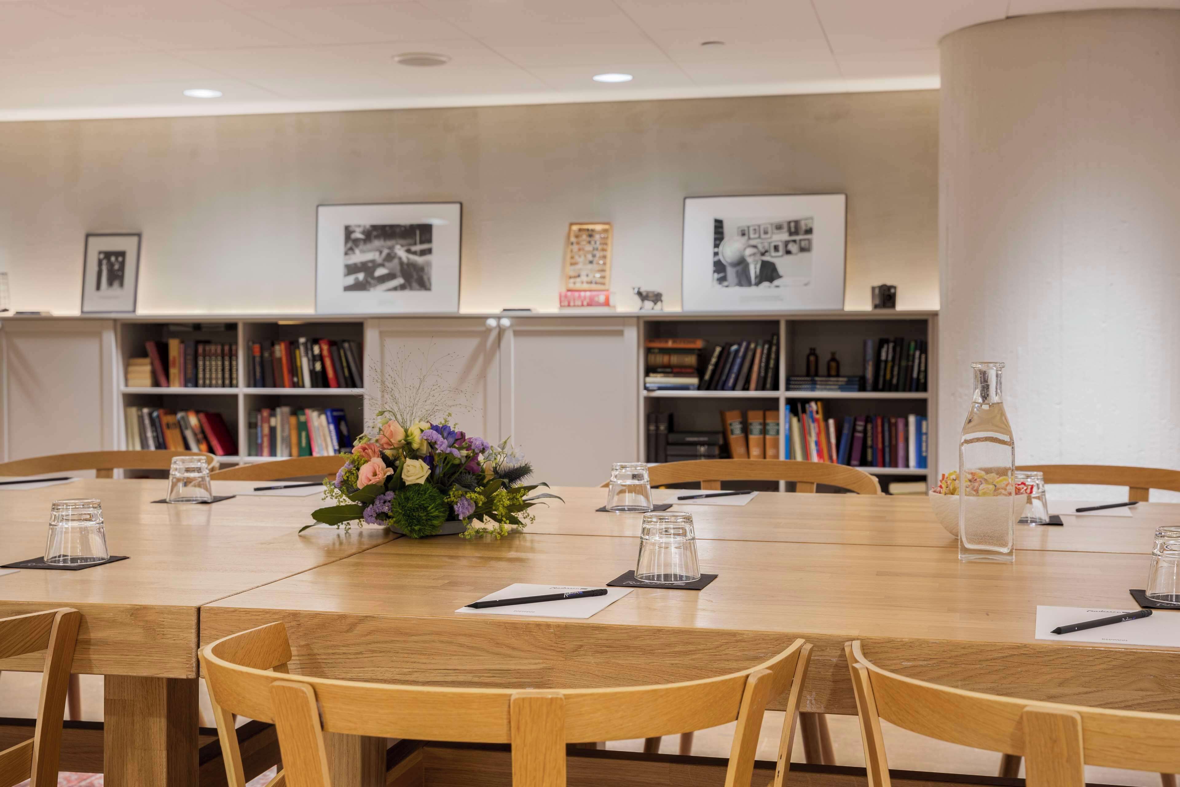A conference room with a wooden table, chairs, a vase of flowers, and bookshelves in the background.