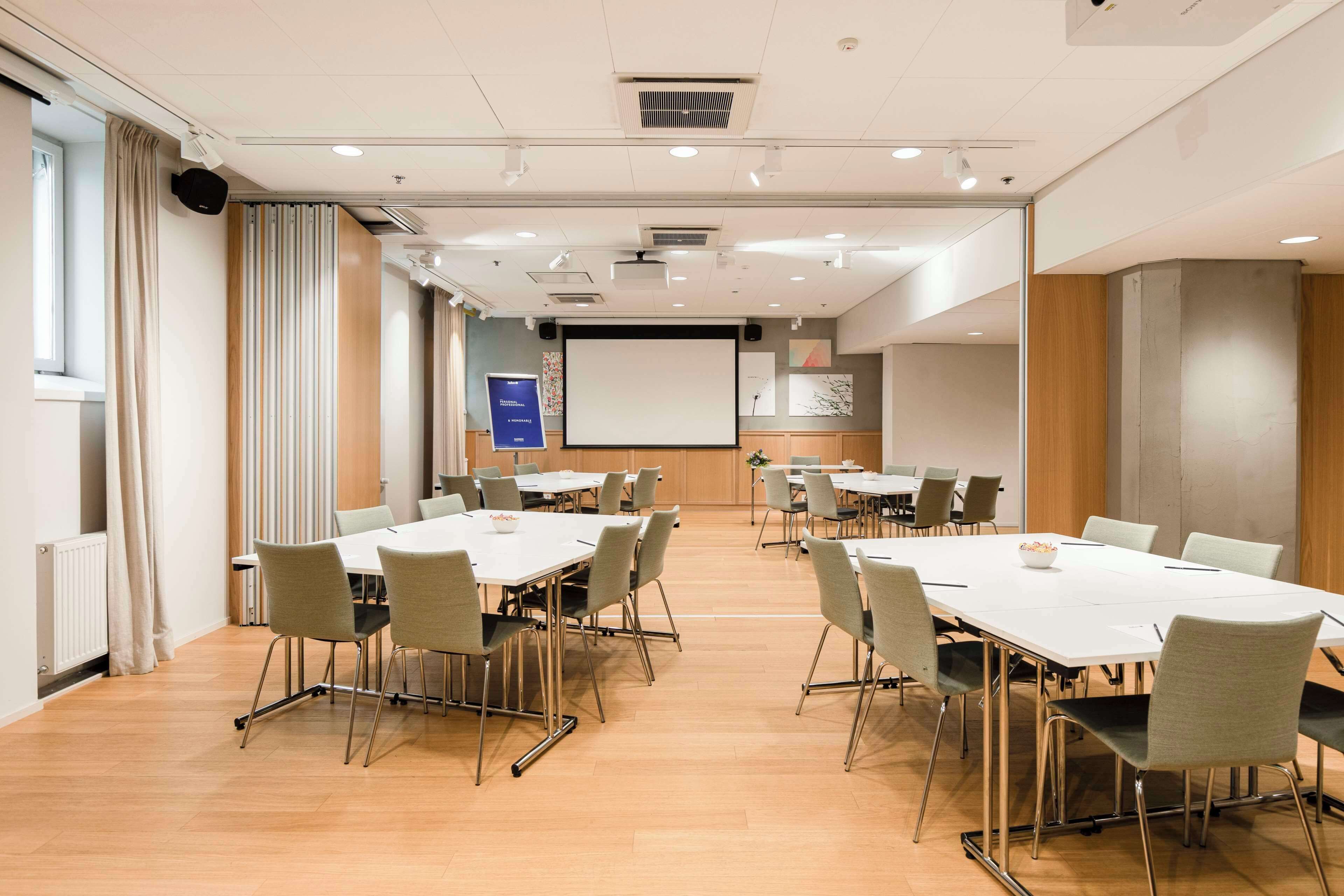 Un salón de conferencias con varias mesas y sillas, una pantalla de proyector y una pizarra para presentaciones.