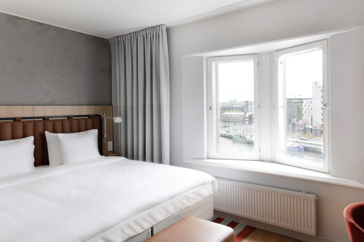 Moderni hotellihuone, jossa on suuri sänky, ikkunasta avautuva näkymä kaupunkiin ja punainen tuoli.