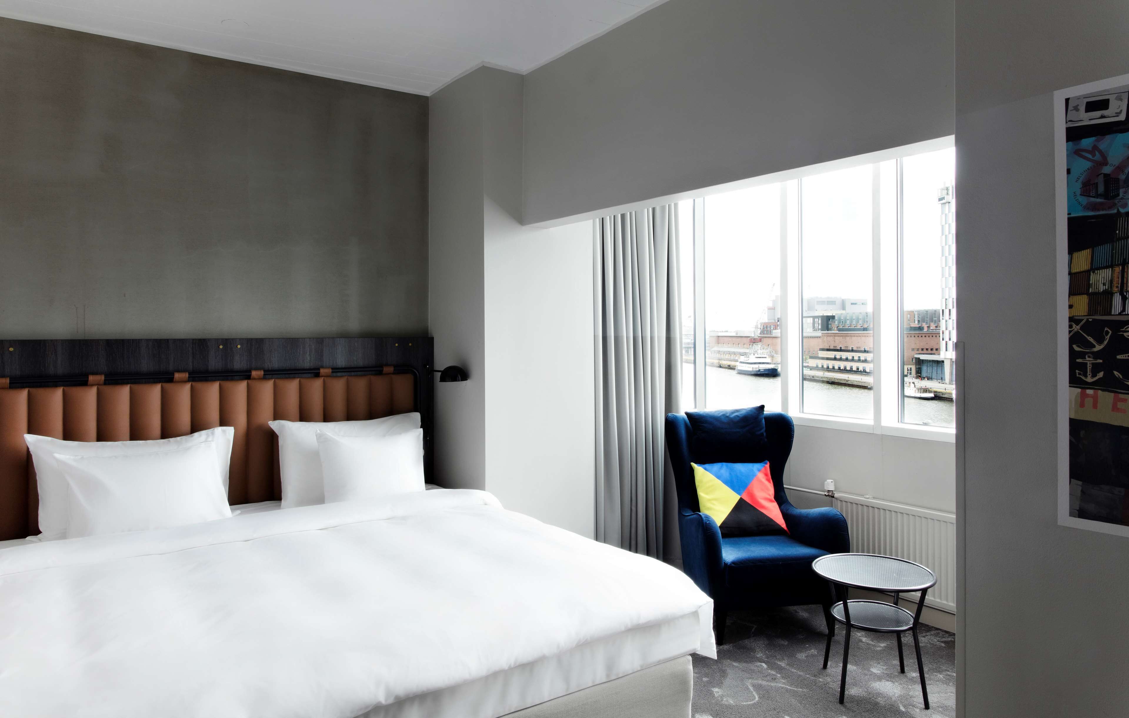 A hotel room with a large bed, a chair, a small table, and a window with a view of buildings.