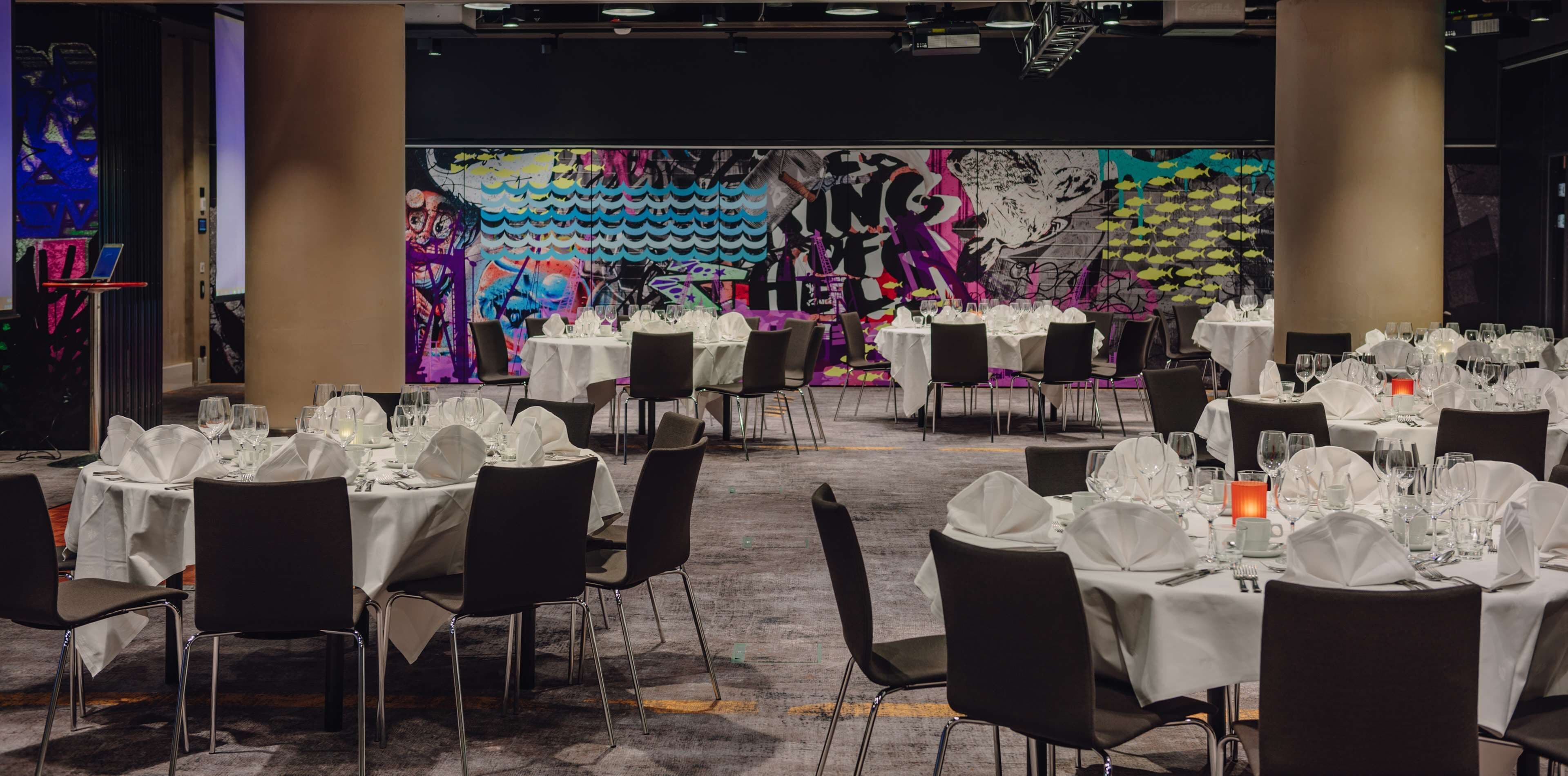 A banquet hall with round tables set for dining, a large abstract mural on the back wall, and a modern chandelier.