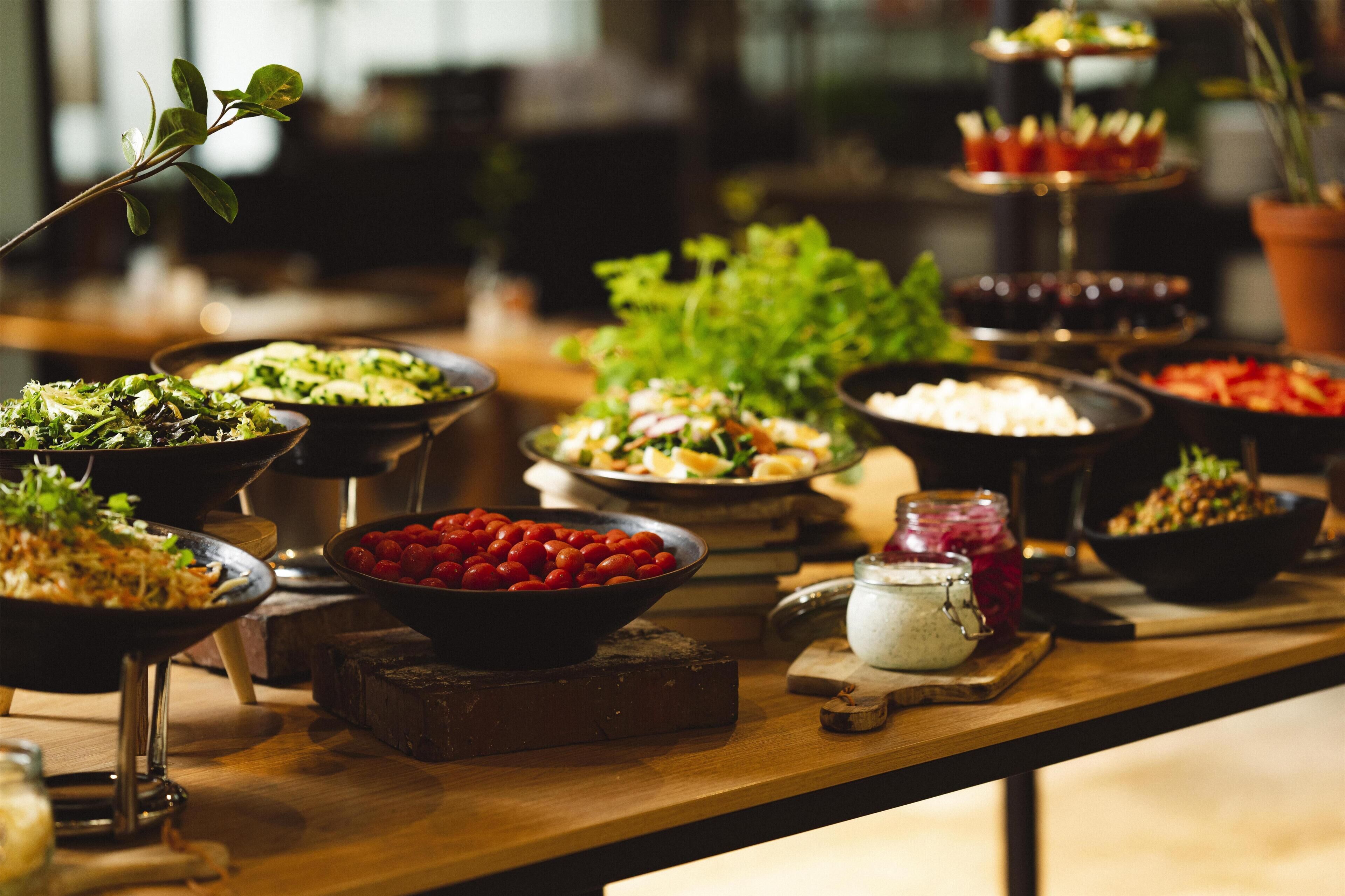 Uma mesa de buffet com diversos pratos, incluindo saladas, legumes e um recipiente com tomates cereja.