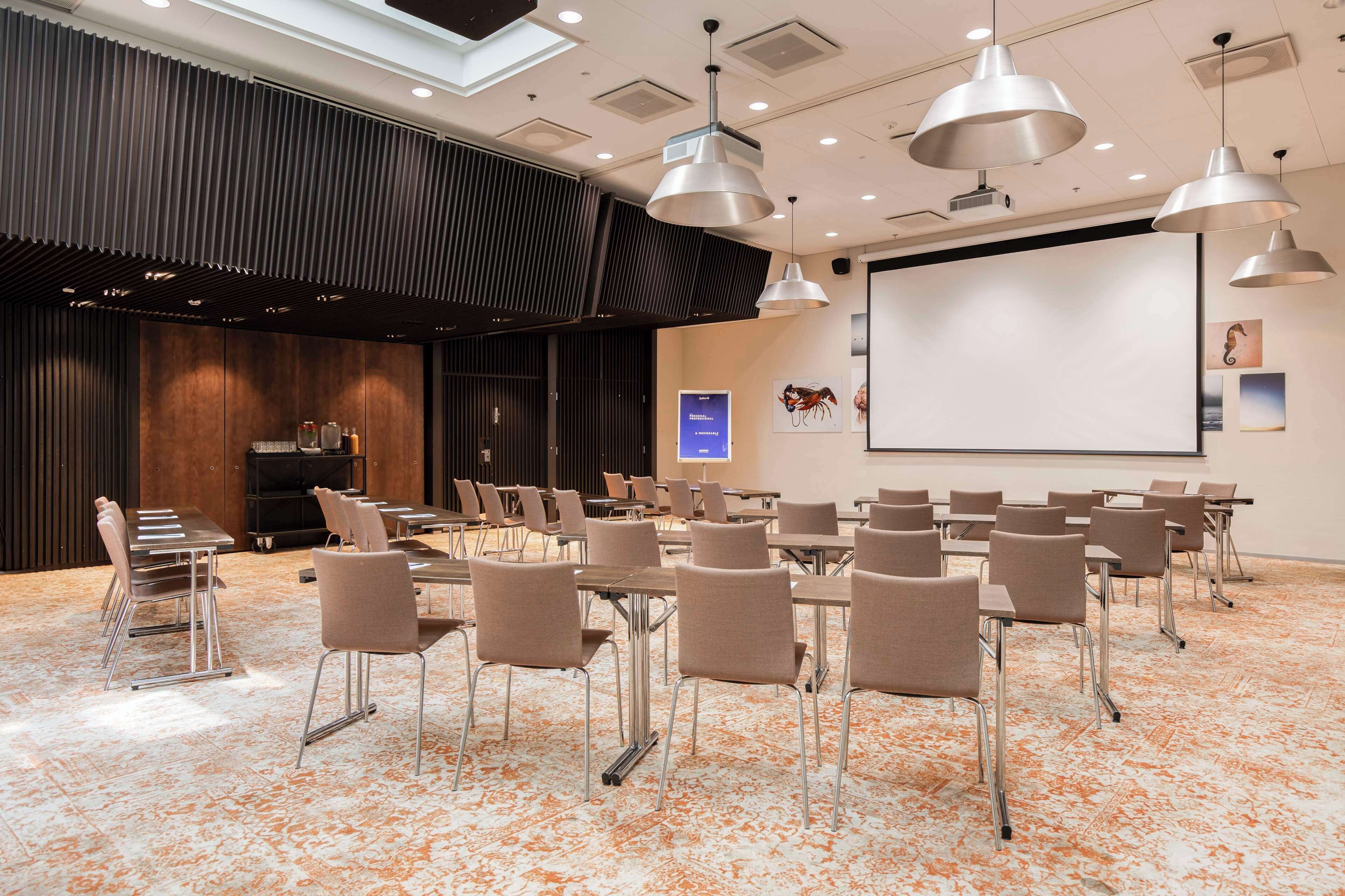 Un salón de conferencias con una pantalla grande, varias mesas y sillas dispuestas para presentaciones.