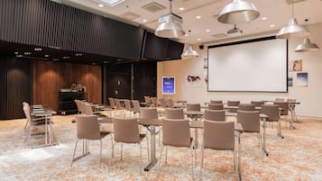 Un salón de conferencias con una pantalla grande, varias mesas y sillas dispuestas para presentaciones.