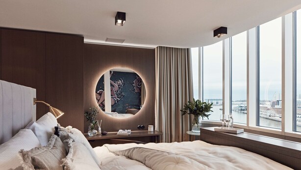 A modern hotel room with a large bed, a round mirror with a cityscape artwork, a view of the city through large windows, and a small table with a vase and glasses. - Radisson Blu Seaside Hotel, Helsinki (Helsinki)