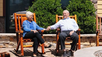 Two people seated on wooden rocking chairs outdoors, with stone walls and green bushes in the background.