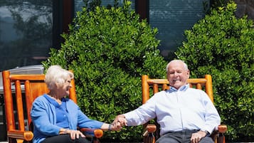 Dos personas sentadas en mecedoras al aire libre, con un edificio y follaje al fondo.