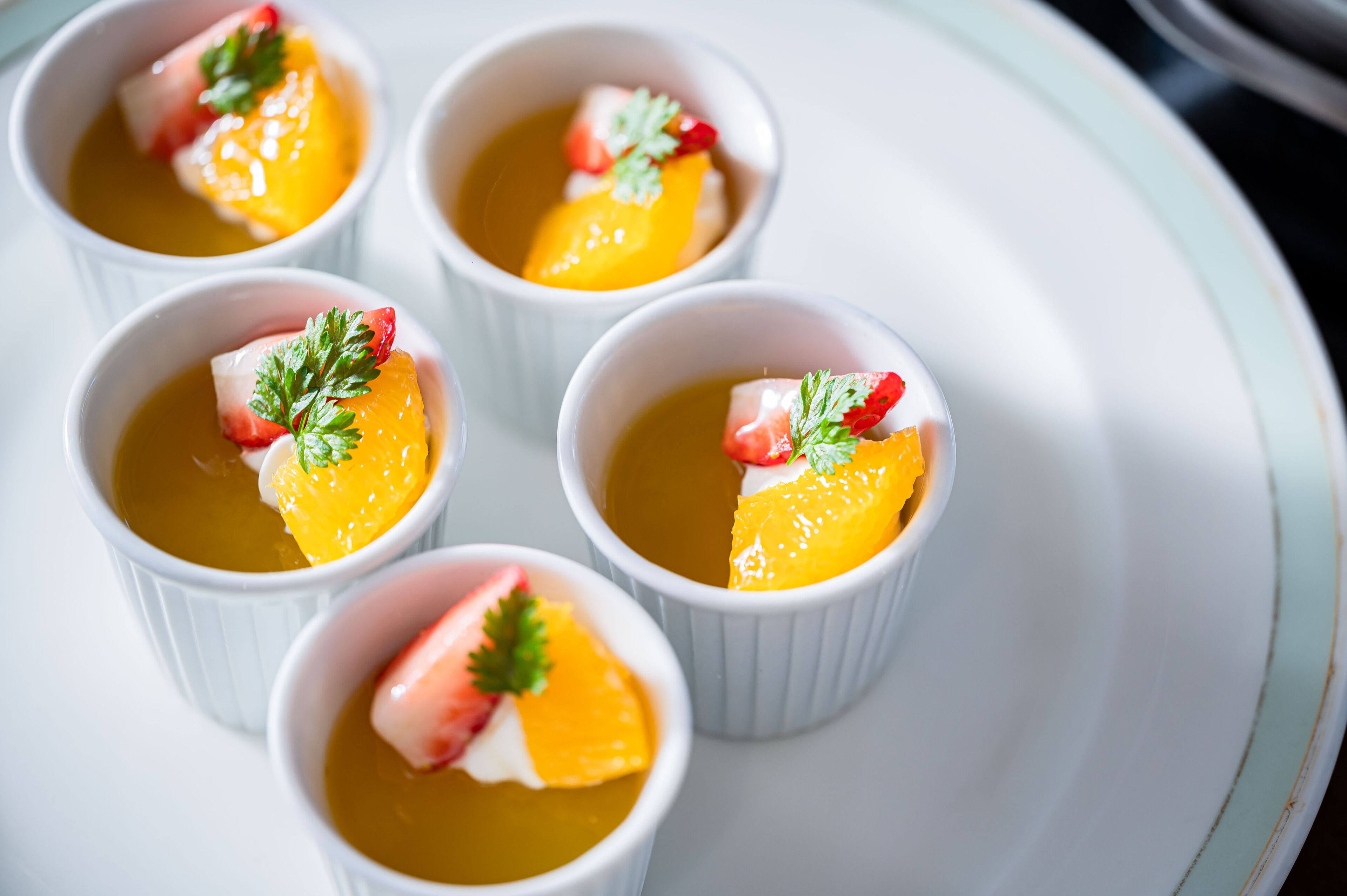 Quatro pequenas tigelas brancas com uma sobremesa amarela, decorada com morangos e salsa, sobre um prato branco.