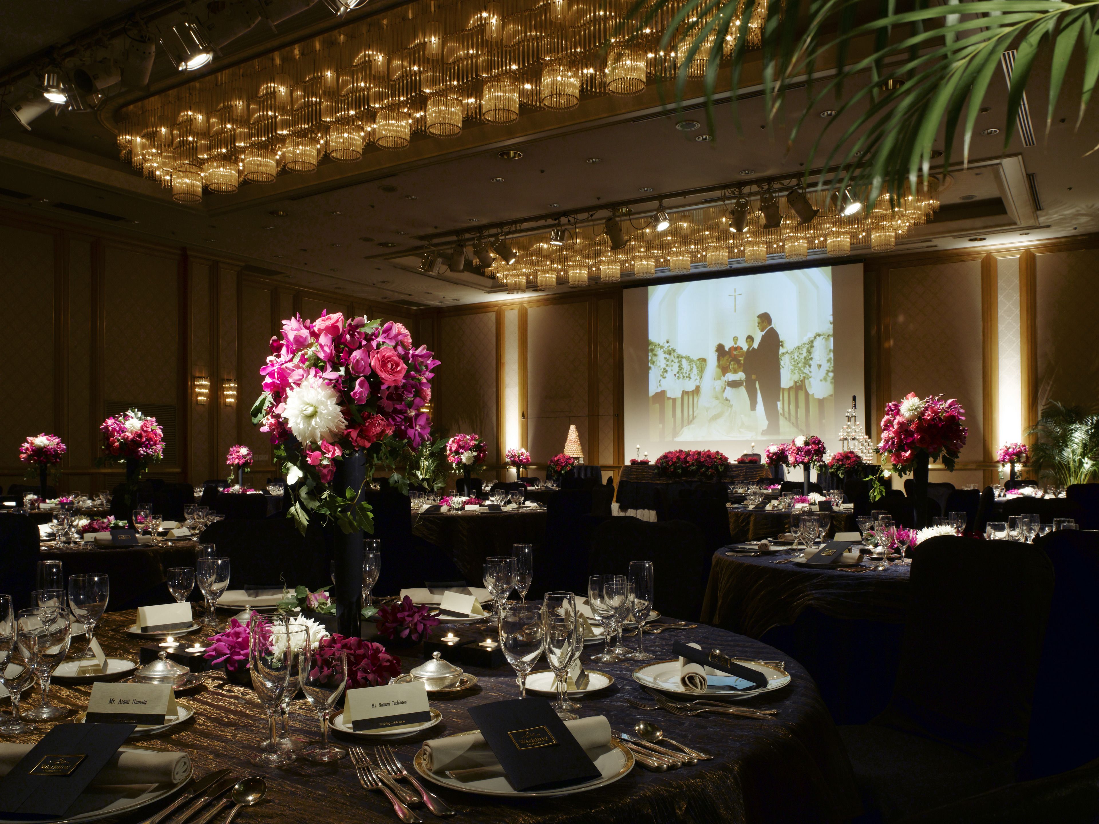 Salão de banquetes com mesas montadas para um evento formal, com arranjos florais no centro e uma tela de projeção ao fundo.